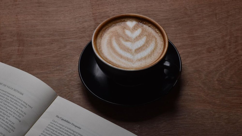 Cappuccino s latte art v černém šálku vedle rozečtené knihy na dřevěném stole